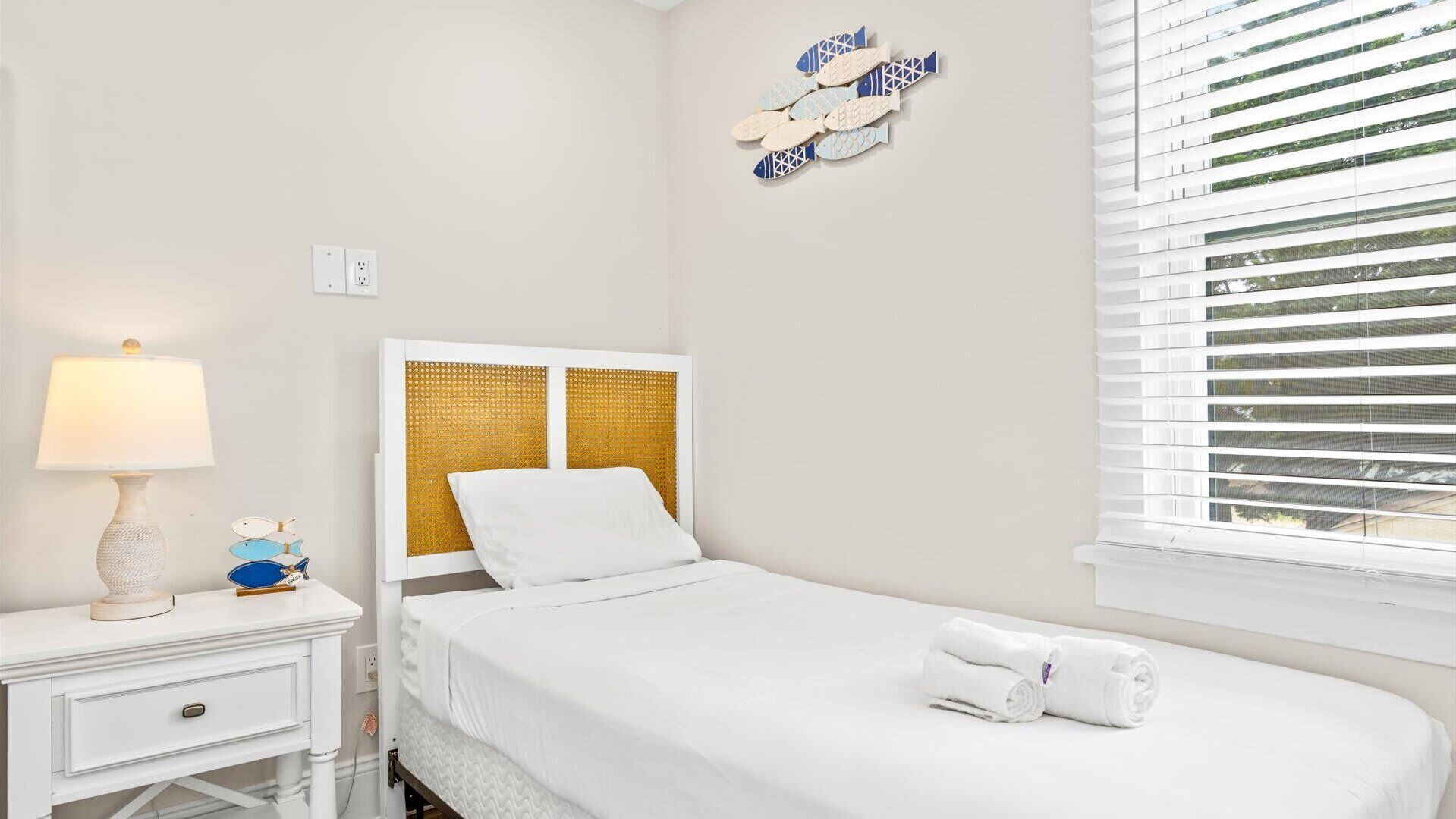 A neatly arranged hotel room with a bed, a bedside table with a lamp, a wall decoration, and a window with blinds.