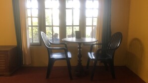 A room with a round table and two chairs, large windows with curtains, and a red carpeted floor. - Manikkal Bungalow Uphill Cottage Large (Mananthavady)