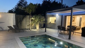 A swimming pool with a tiled deck, lounge chairs, a dining area with chairs and a table, and a covered patio area with blue LED lights.