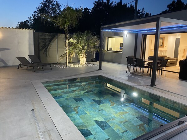 Une piscine avec une terrasse carrelée, des chaises longues, un coin repas avec des chaises et une table, et un patio couvert agrémenté de lumières LED bleues.