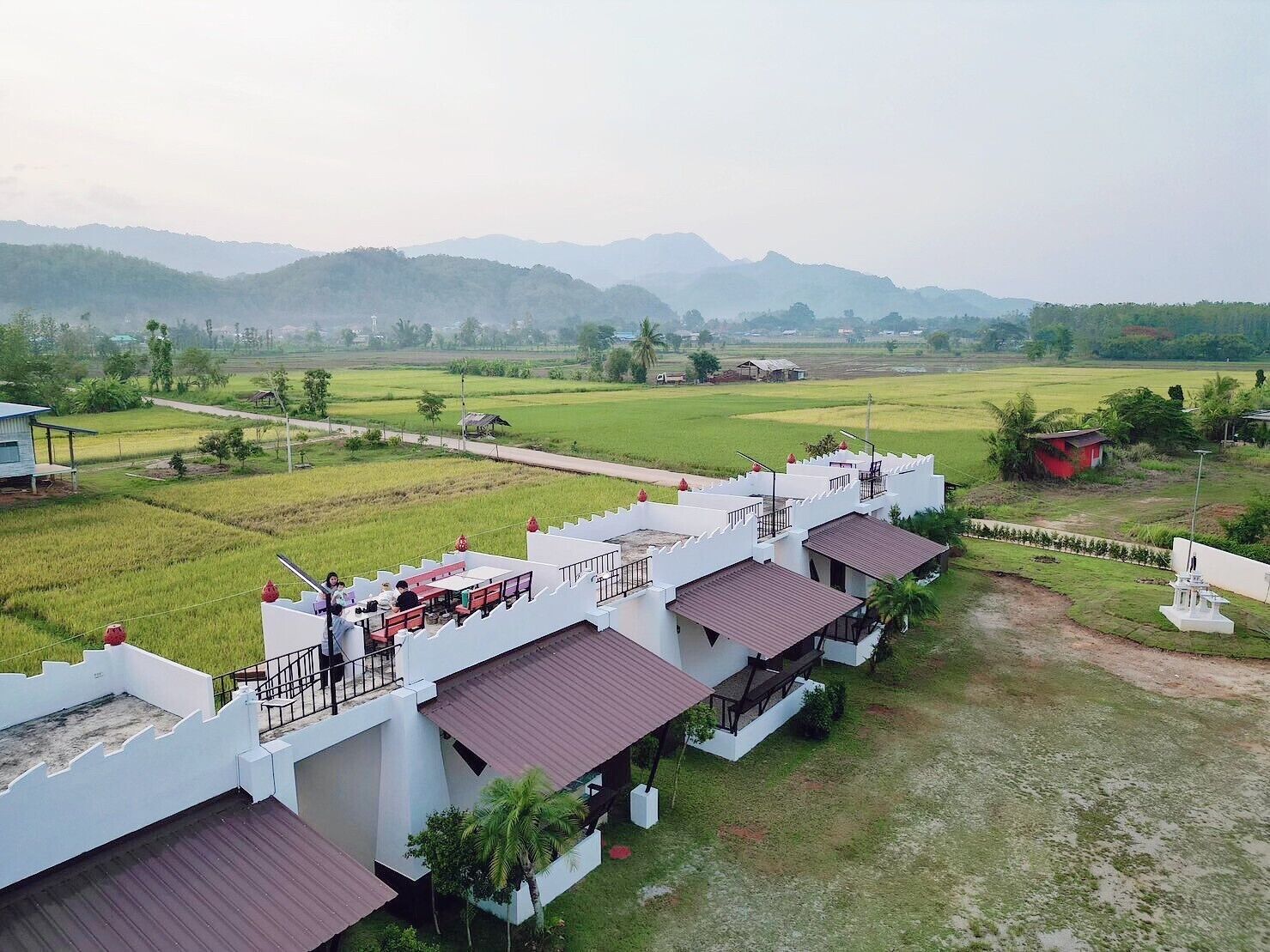 A hotel with multiple white buildings, each with a red roof and a balcony. The surrounding area includes green fields and distant mountains.