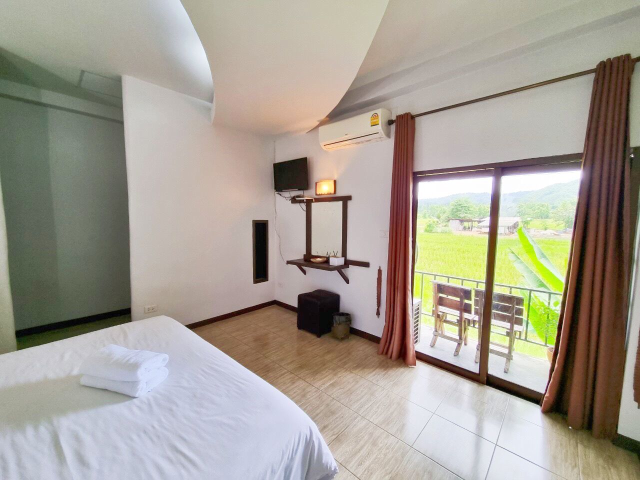 Habitación de hotel con cama, televisor, mesita con lámpara, cubo de basura y balcón con vistas al campo.
