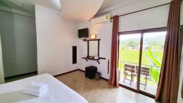 Habitación de hotel con cama, televisor, mesita con lámpara, cubo de basura y balcón con vistas al campo.