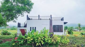 A white two-story house with a balcony, surrounded by lush greenery and a body of water.