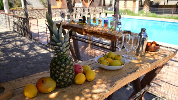 Una mesa de madera con frutas, vasos y botellas cerca de una piscina.
