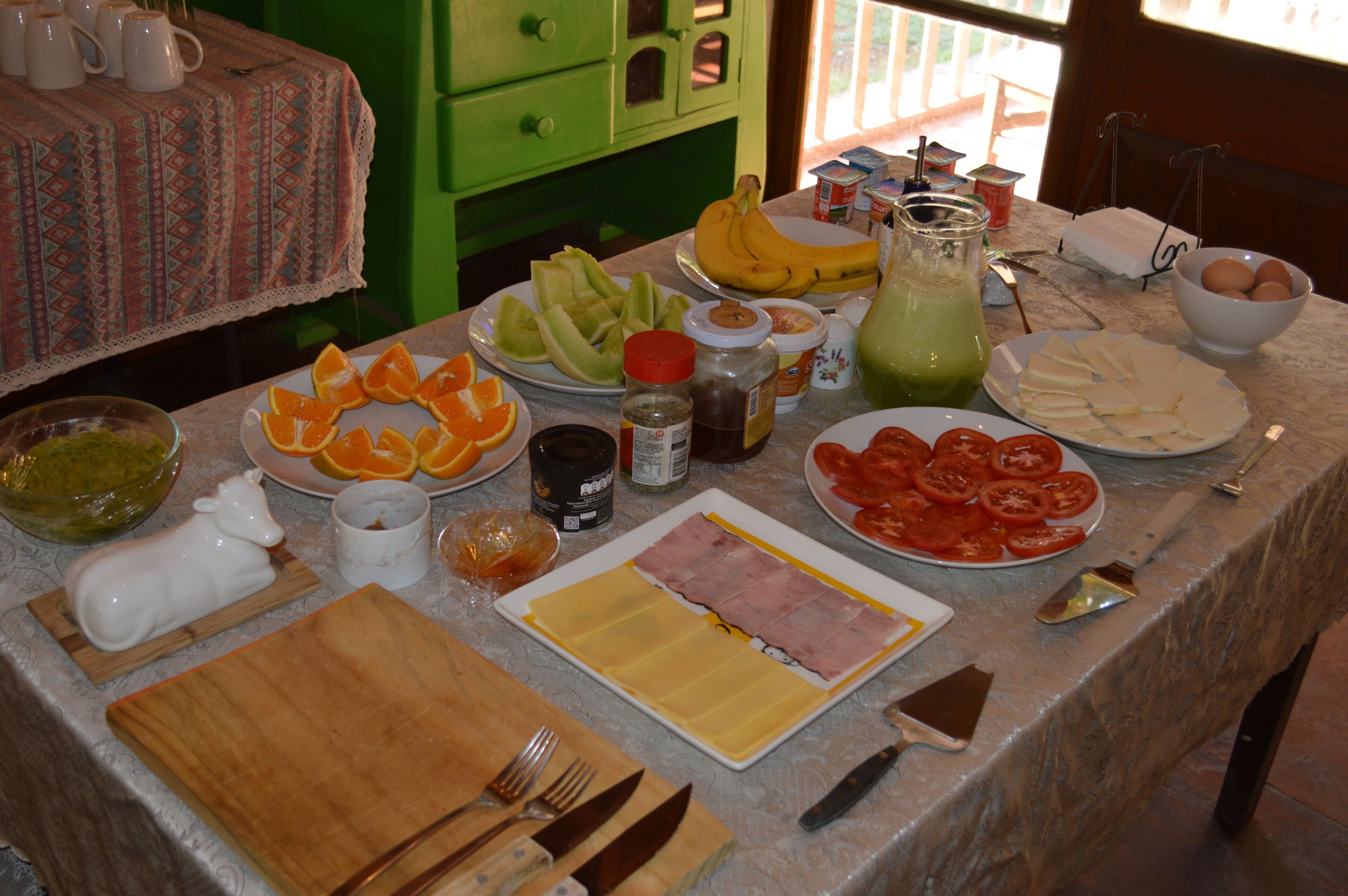 Una mesa con diversos alimentos y condimentos, como platos de tomates en rodajas, naranjas y queso, además de una jarra de vidrio con jugo verde.