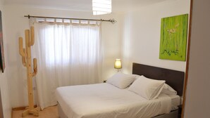 Una cama bien hecha con ropa blanca, piso de madera, una lámpara de pared y un cuadro de flores.