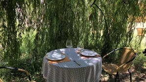 Una mesa redonda con mantel de encaje, dos platos y una copa de vino, al aire libre, rodeada de sillas de mimbre y un sauce llorón.