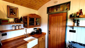 A kitchen with wooden cabinets, a built-in oven, a coffee maker, and a shelf with various kitchen items. - Exquisite Shepherd's HUT With HOT TUB - Between Moor and sea (Camelford)