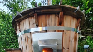 A wooden outdoor sauna with a visible fire burning inside, surrounded by gravel and logs. - Exquisite Shepherd's HUT With HOT TUB - Between Moor and sea (Camelford)