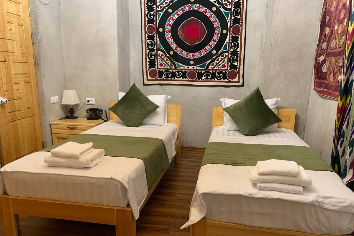A room with two single beds, each with a green pillow and a white towel. There is a wall tapestry above the beds, a wooden door to the left, and a small table with a lamp and phone.