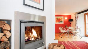 A modern living room with a wood-burning stove, a dining area with a red wall and a painting, and a wicker basket with logs. - Great Brightor Cob Barn (Bodmin)