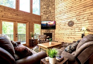 A cozy living room with leather sofas, a stone fireplace, a television mounted on the wall, and a large window overlooking greenery. - Yourcation® Awaits in Spectacular, 6-Bedroom Cabin! (Sevierville)