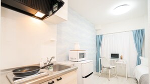 Uma cozinha compacta com micro-ondas, fogão e pia. Há uma pequena mesa com um laptop e uma cadeira. O quarto tem cortinas azul claro e uma cama branca.