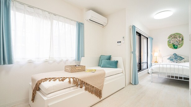 Quarto com sofá branco, cama com estrutura branca, janela com cortinas azuis e unidade de ar condicionado na parede.