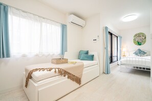 A bedroom with a white sofa, a bed with a white frame, a window with blue curtains, and an air conditioning unit on the wall. - ZAITO Tokyo Luna Lane Economy Inn (Tokyo)