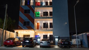 Un edificio de varios pisos con un letrero que dice 'J's Way', múltiples balcones y varios coches estacionados al frente.