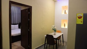 Una habitación pequeña con una cama, una mesa con un florero y un estante en la pared con una lámpara.