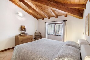 A bedroom with a wooden ceiling, a bed with patterned bedding, a dresser, a chair, and a window with curtains.
