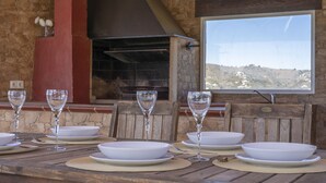 Une table à manger dressée avec de l’argenterie blanche et des verres, un mur en pierre et, par la fenêtre, une vue sur une colline.