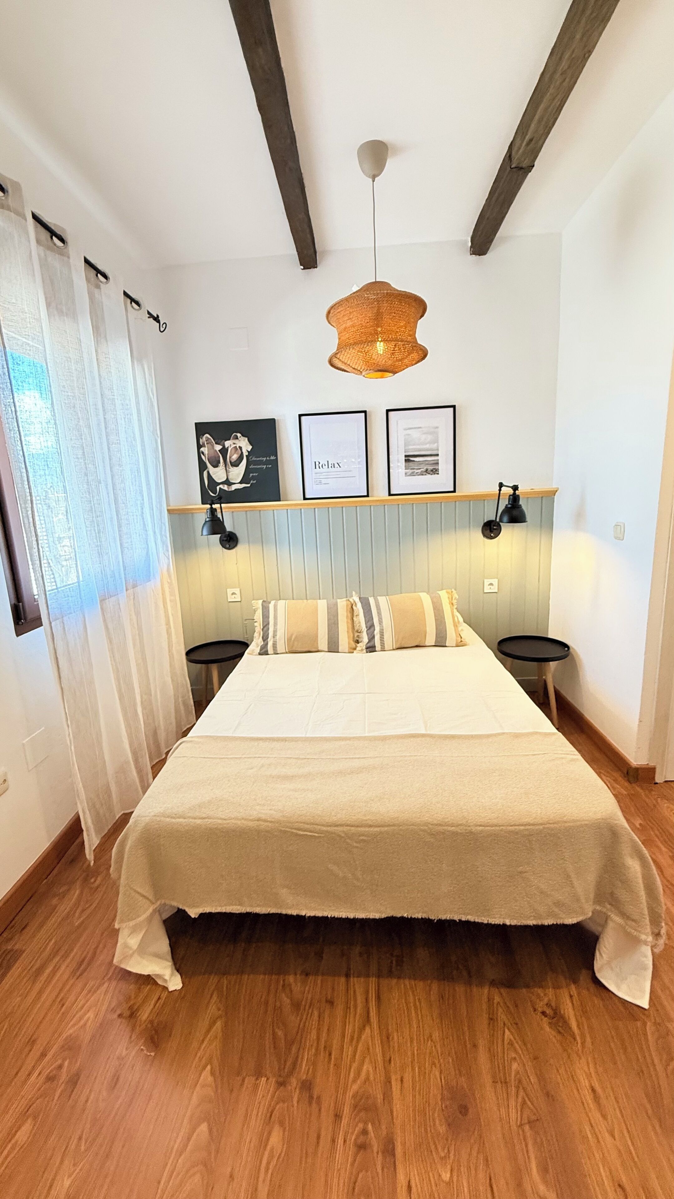 A bedroom with a large bed, wooden flooring, a hanging light fixture, and framed pictures on the wall.