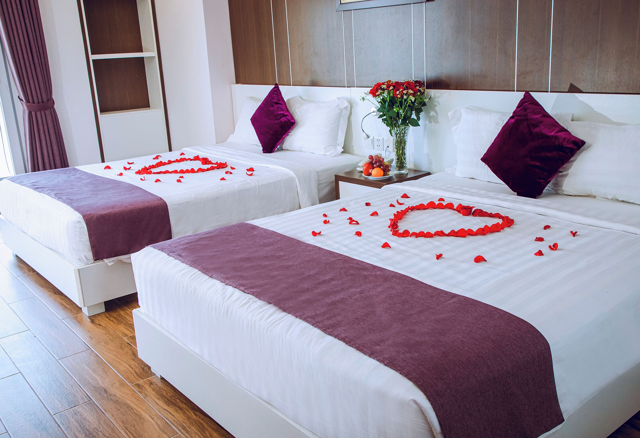 Two beds with white linens, red rose petals on the beds, and a vase with flowers on a nightstand.