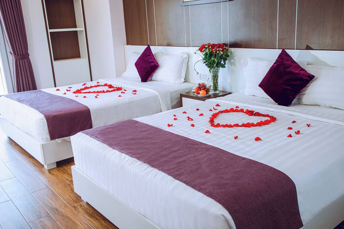 Two beds with white linens, red rose petals on the beds, and a vase with flowers on a nightstand.