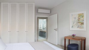 Un dormitorio con cama, una mesita de noche de madera, un armario blanco y un baño visible a través de una puerta abierta.
