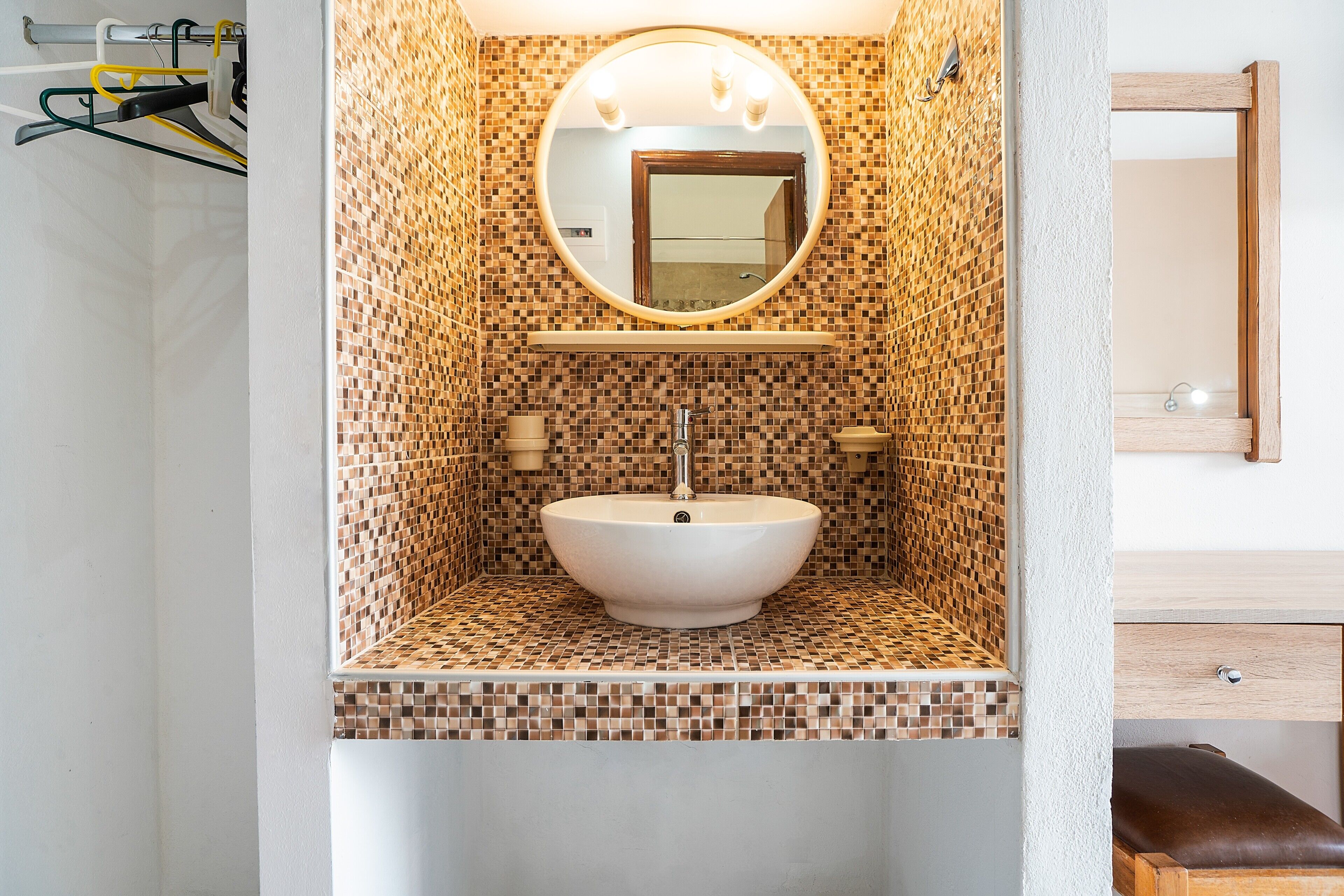 Ein Badezimmer mit einer mosaikbesetzten Wand, einem runden Spiegel, einem weißen Waschbecken und einer hölzernen Sitzbank.