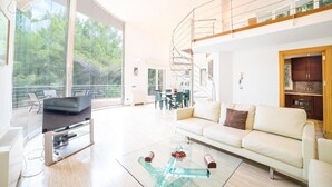 Een ruime woonkamer met een bank, salontafel en televisie. Een trap leidt naar een verdieping hoger, en een balkon met uitzicht op bomen.