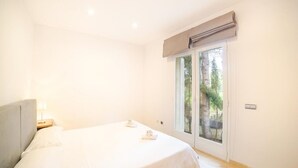 Een slaapkamer met een bed, nachtkastje, lamp en raam met uitzicht op bomen.