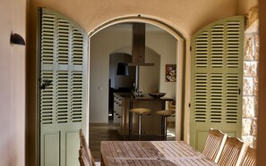 Uma área de refeições com uma mesa e cadeiras de madeira, uma cozinha ao fundo e portadas verdes nas paredes.