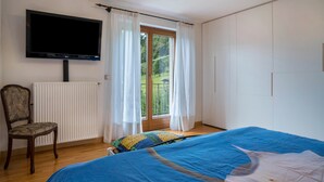 Een slaapkamer met een bed, een stoel, een televisie, een raam met gordijnen en uitzicht op een balkon.
