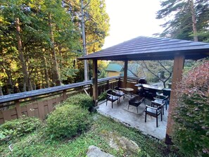Ein überdachter Patio-Bereich mit Gartenmöbeln, einem Grill und Blick auf einen bewaldeten Bereich.