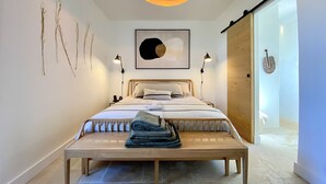 A bedroom with a bed, bedside lamps, a hanging light fixture, a wall-mounted artwork, and a bench with towels.