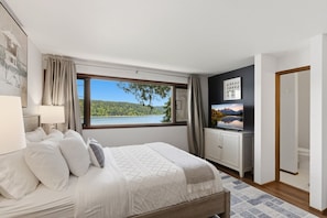 A bedroom with a bed, a TV, a window with a view of a lake, and a door.