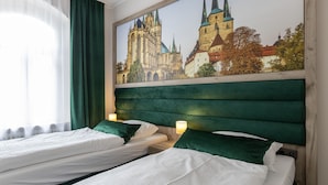 Een hotelkamer met twee bedden, groene hoofdeinden, witte beddengoed en een groot raam met dunne gordijnen.