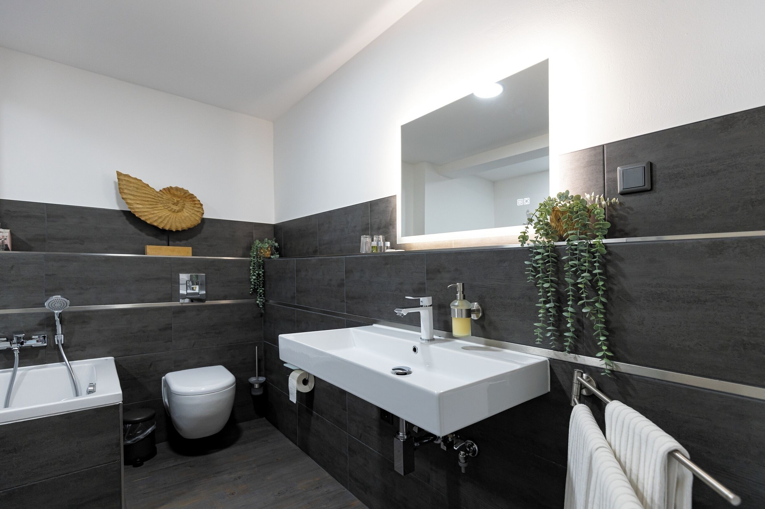 Un baño moderno con un lavabo blanco, un inodoro y una bañera. Hay un espejo grande sobre el lavabo y una hoja decorativa en la pared.