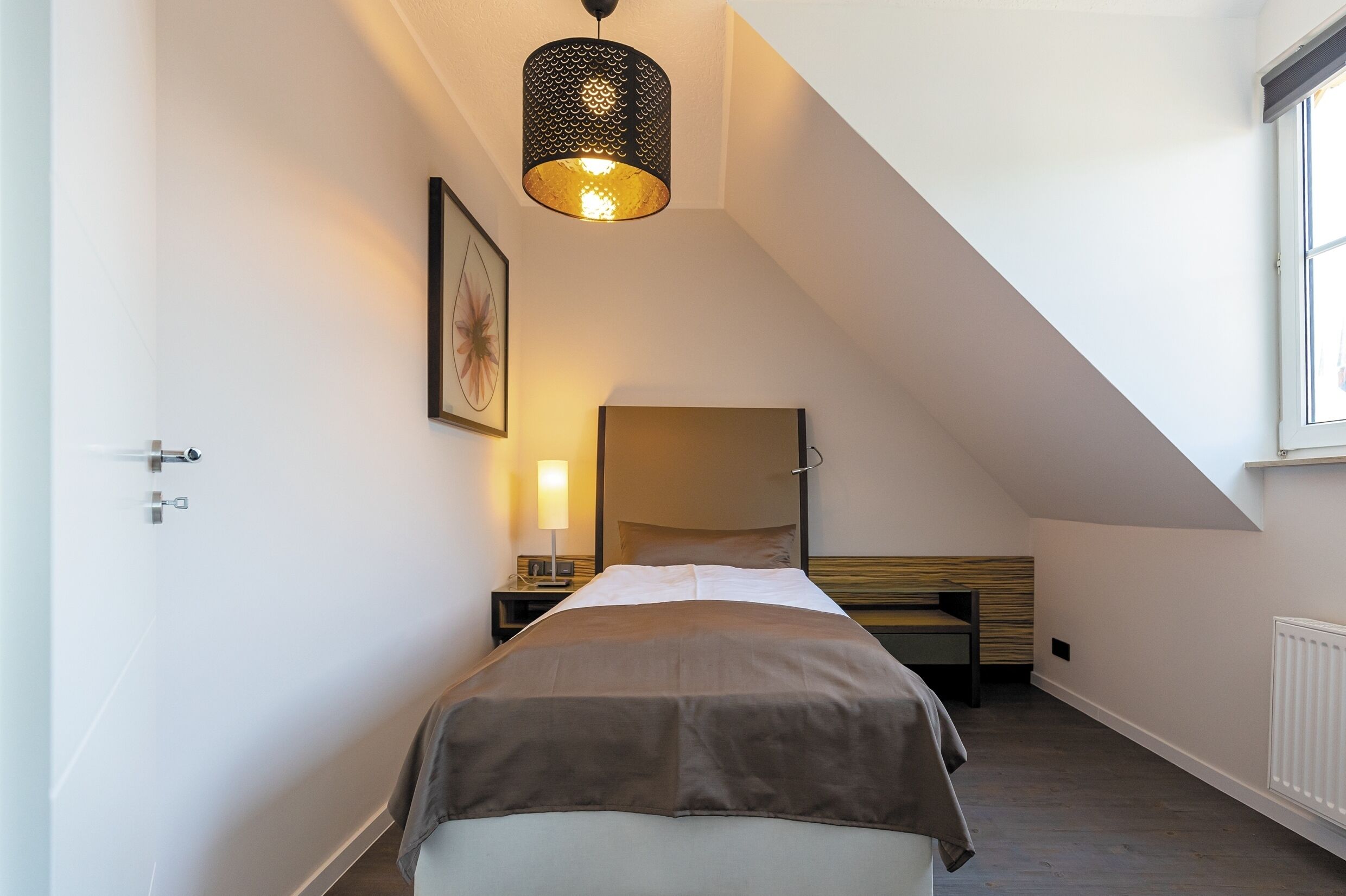 A bedroom with a sloped ceiling, a bed with a brown bedspread, a bedside lamp, a framed picture on the wall, and a window on the right.