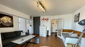 A modern living room with a sofa, coffee table, and a dining area. There is a kitchen in the background, a door leading to another room, and a lamp on a side table. - Port Apartment 2 Rooms Saint-Cyprien with Parking (Saint-Cyprien)
