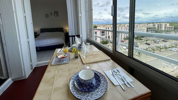 A hotel room with a balcony, a bed, a dining table set with plates, a cup, and cutlery. - Port Apartment 2 Rooms Saint-Cyprien with Parking (Saint-Cyprien)