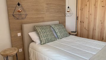 Un dormitorio con una cama, dos almohadas estampadas, un cabecero de madera, dos lámparas colgantes, una mesita redonda pequeña y una puerta de madera.