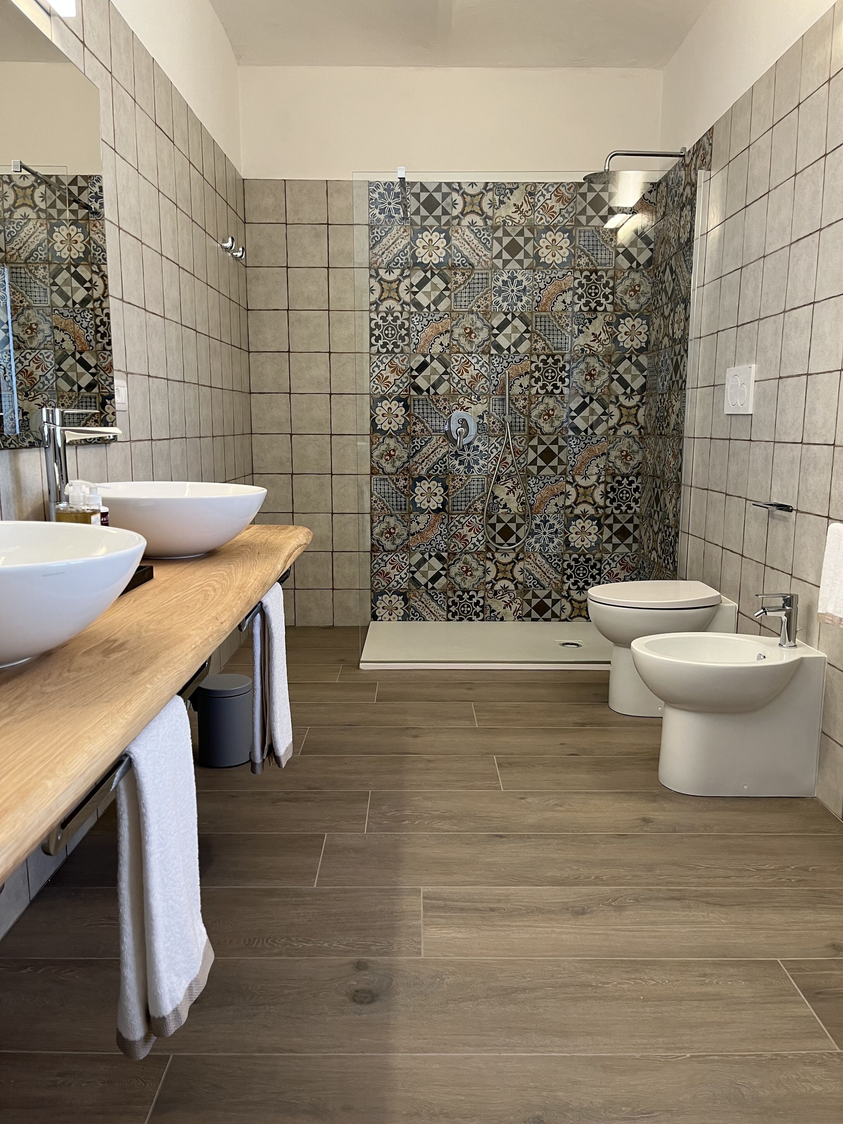 A modern bathroom with a wooden vanity, two sinks, a toilet, and a shower area with patterned tiles.