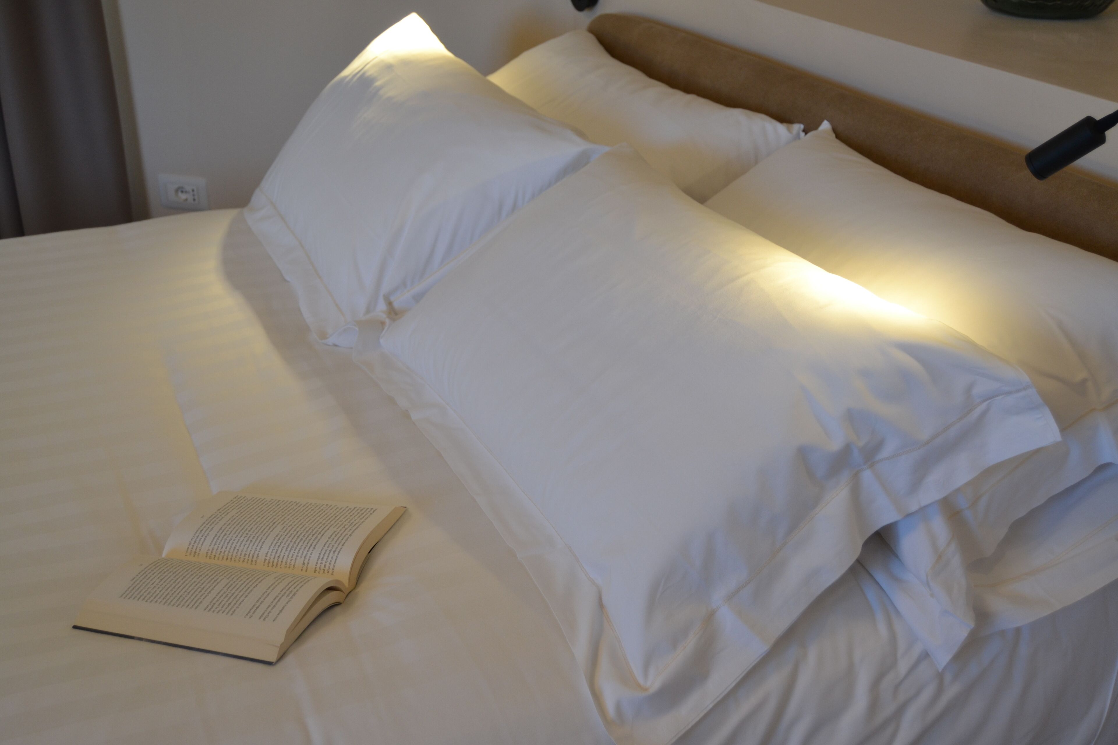 Una cama bien tendida con sábanas y almohadas blancas, un libro sobre la cama y una lámpara fijada a la pared.