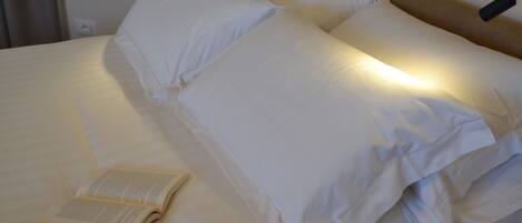 A neatly made bed with white linens and pillows, a book on the bed, and a wall-mounted lamp.