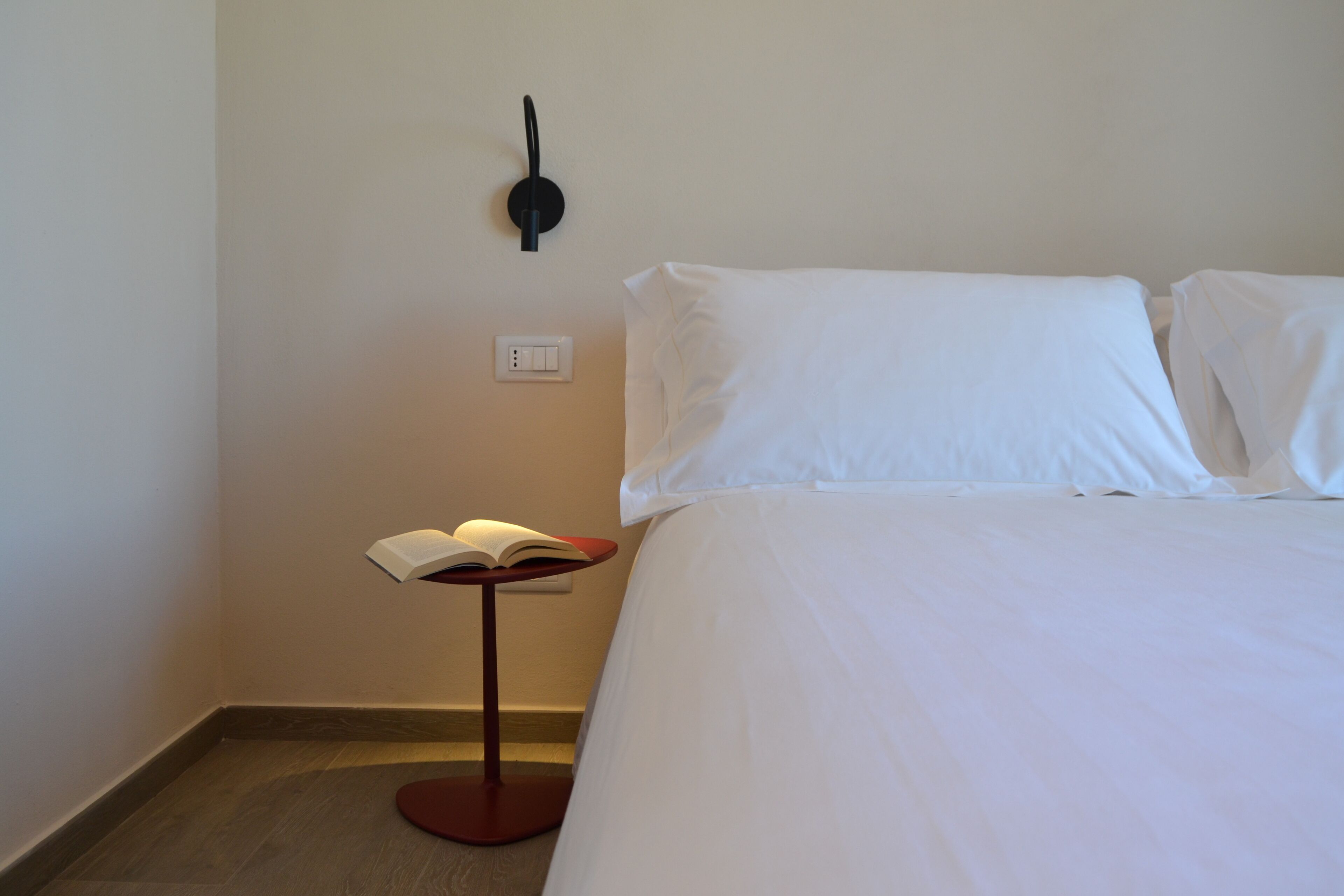 A hotel room with a neatly made bed, a bedside table with a book, a wall-mounted light, and a power outlet.