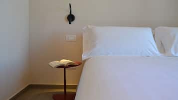 Quarto de hotel com cama bem arrumada, mesa de cabeceira com um livro, luminária na parede e tomada.