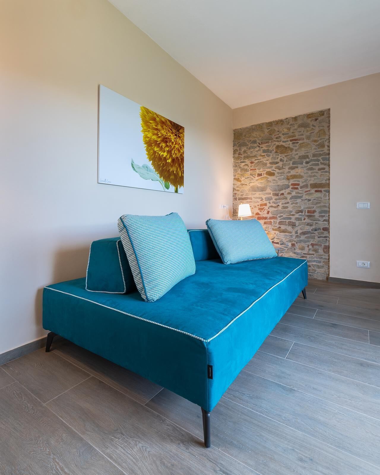 Un sofá cama color turquesa con almohadas a juego, una pared de piedra y un cuadro de una flor en la pared.
