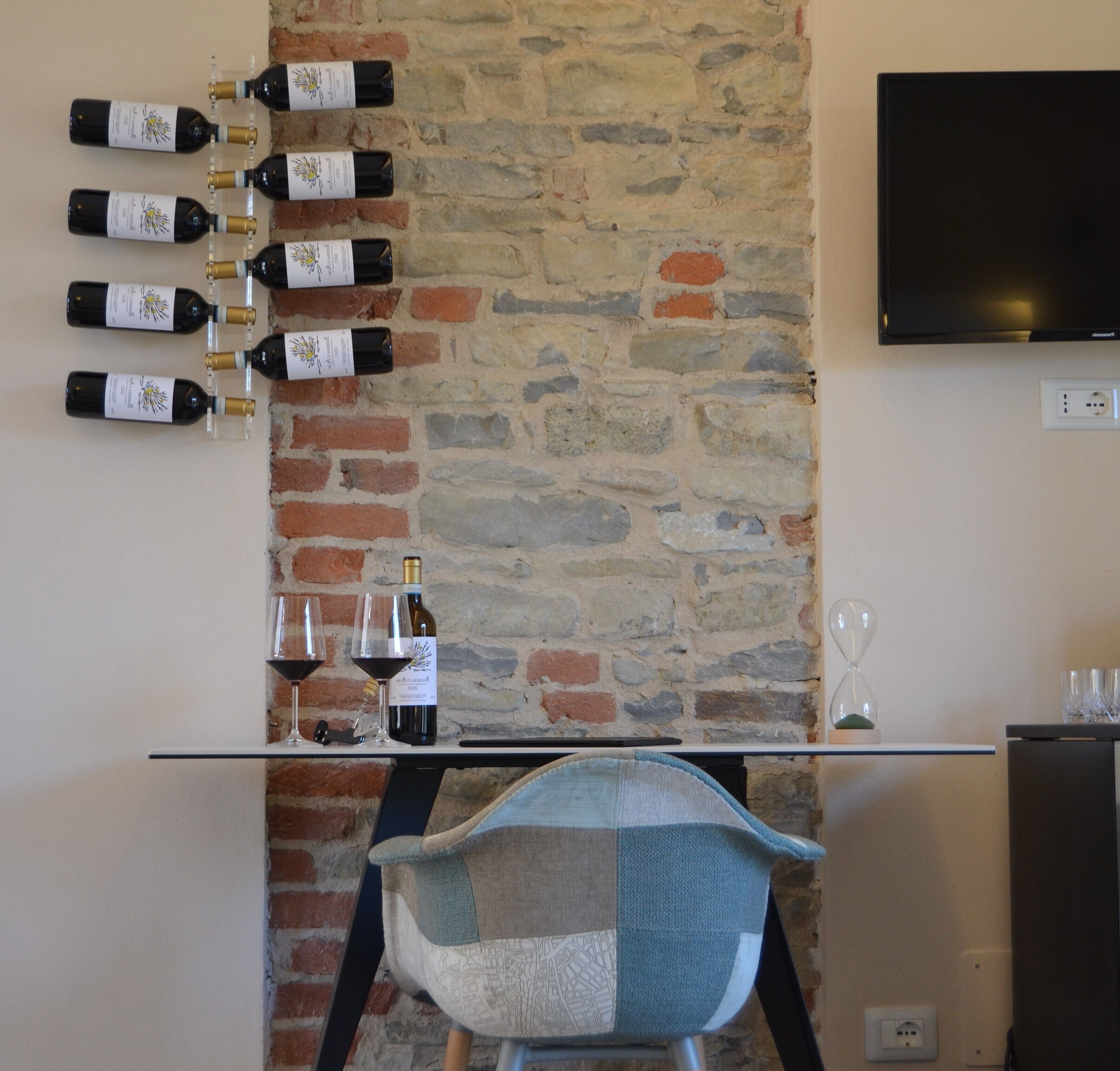 Una habitación con pared de piedra, un televisor de pantalla plana montado en la pared, un botellero con varias botellas, dos copas de vino y una silla con diseño de retazos.
