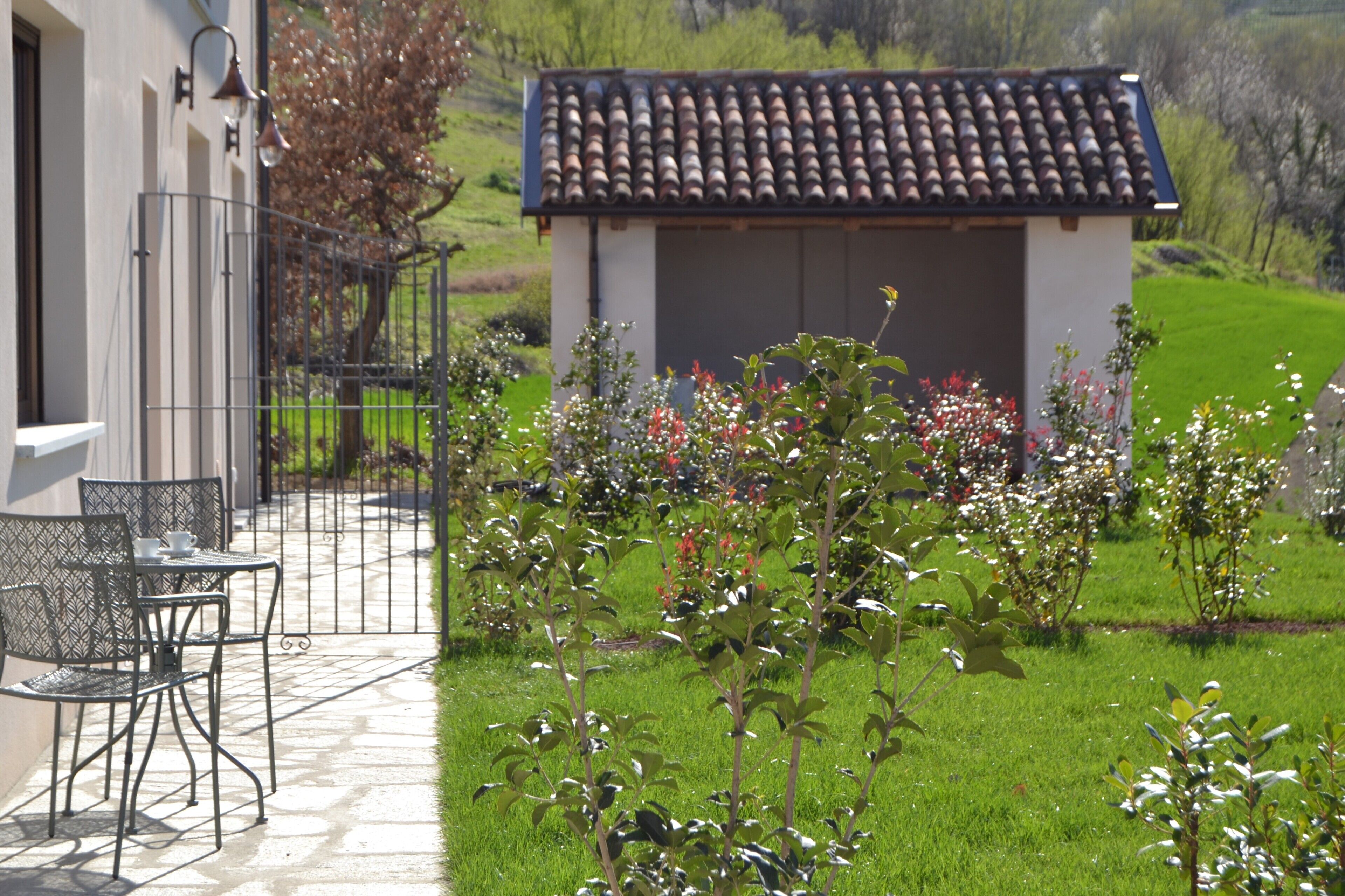 Une terrasse avec des chaises en métal et une petite table, un jardin avec différentes plantes et fleurs, et un bâtiment doté d’un toit en tuiles.